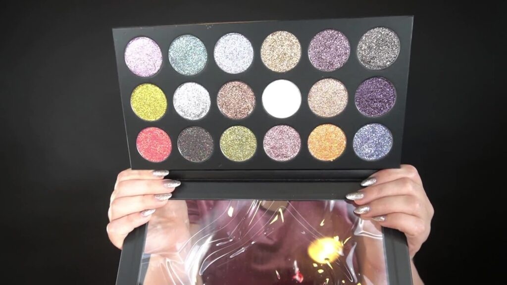 A still frame of the Vani Cosmetics glitter review by Unfiltered Makeup on YouTube is featured. Annmarie Gustafson is pictured holding up the Vani Cosmetics 18 pan pressed glitter palette. The colors are mainly neutrals with a few pops of colors including orange, green, and purple. 